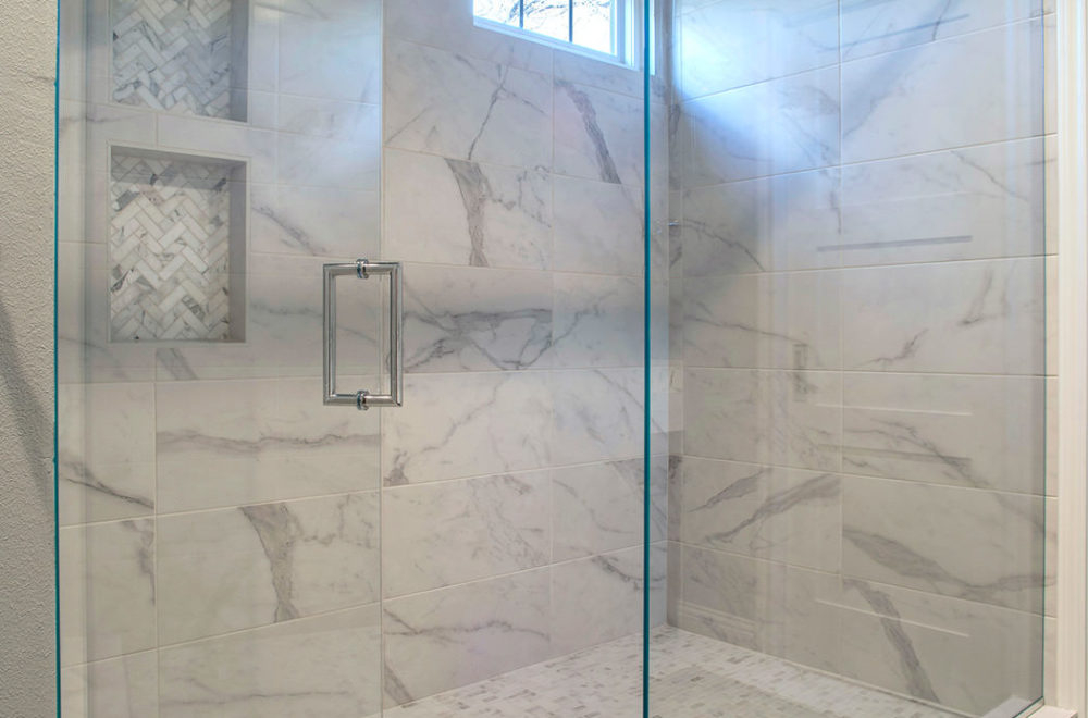 An interior view of a shower featuring white marble tiles on the walls and floor. A herringbone patterned tile accentuates the back wall. The shower is equipped with a glass door, silver handle, and fixtures with a built-in shelf for shower products.