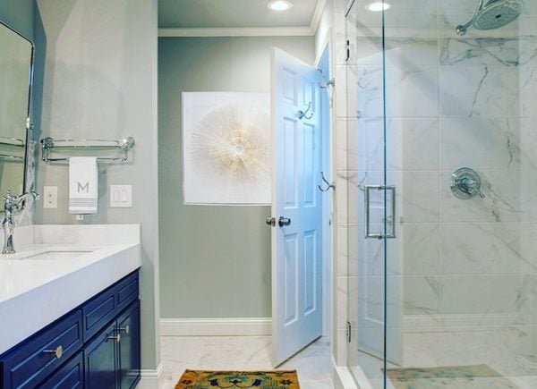 A chic bathroom with a glass-enclosed shower, marble tiles, dark blue vanity, and a light grey wall with decorative art.