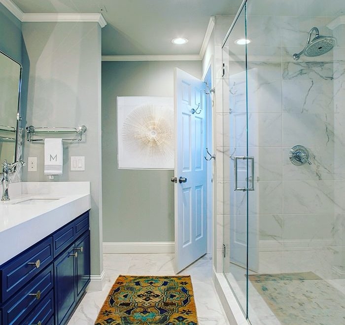 A bright bathroom corner with a walk-in shower enclosed by glass doors. The walls are tiled in a white marble design, and there's a built-in niche for toiletries. A white door and towel hanger are visible, with a light blue artwork on the wall.