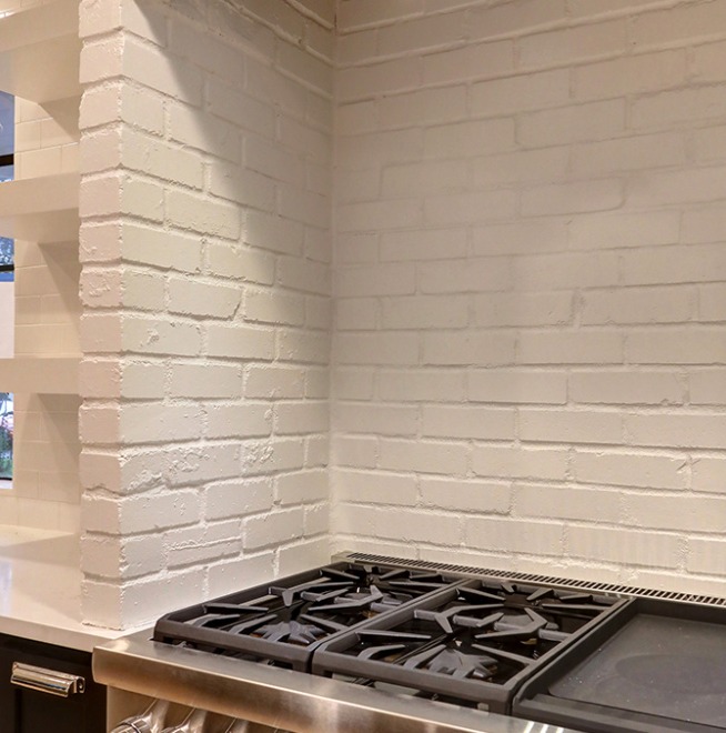 A close-up of a kitchen corner with a white painted brick backsplash and a stainless steel gas stovetop. The countertops are white, and there is a view of the window to the side.