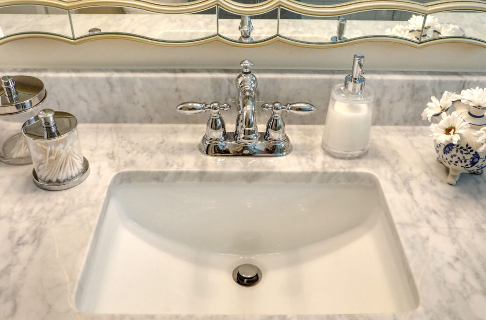 A close-up of a bathroom sink with chrome fixtures. On the marble countertop, there are various bathroom accessories, including a soap dispenser and a container with cotton swabs.