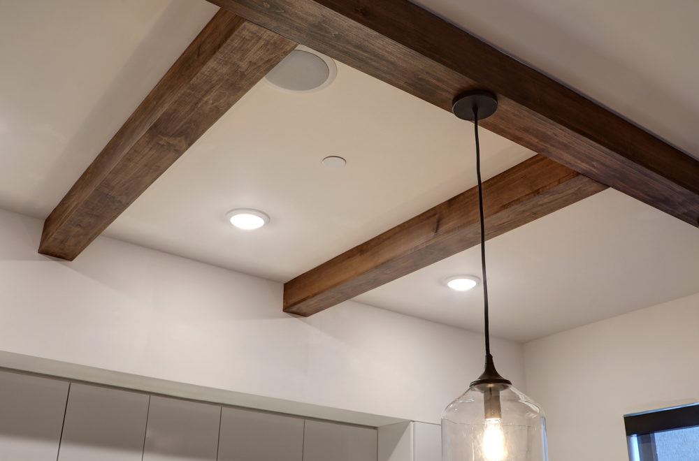 A close-up of a kitchen ceiling showing an architectural detail with wooden beams crossing at an angle, complemented by recessed lighting and a modern hanging light fixture.