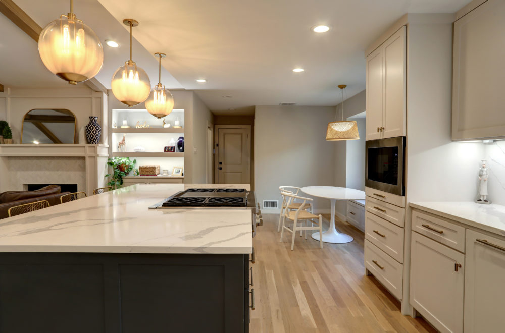 A view of the kitchen from the island looking towards the open-plan living area. The kitchen features white cabinetry, a white countertop, and a set of three globe pendant lights. There are metal bar stools at the island, and the space is well-lit with natural and artificial light.