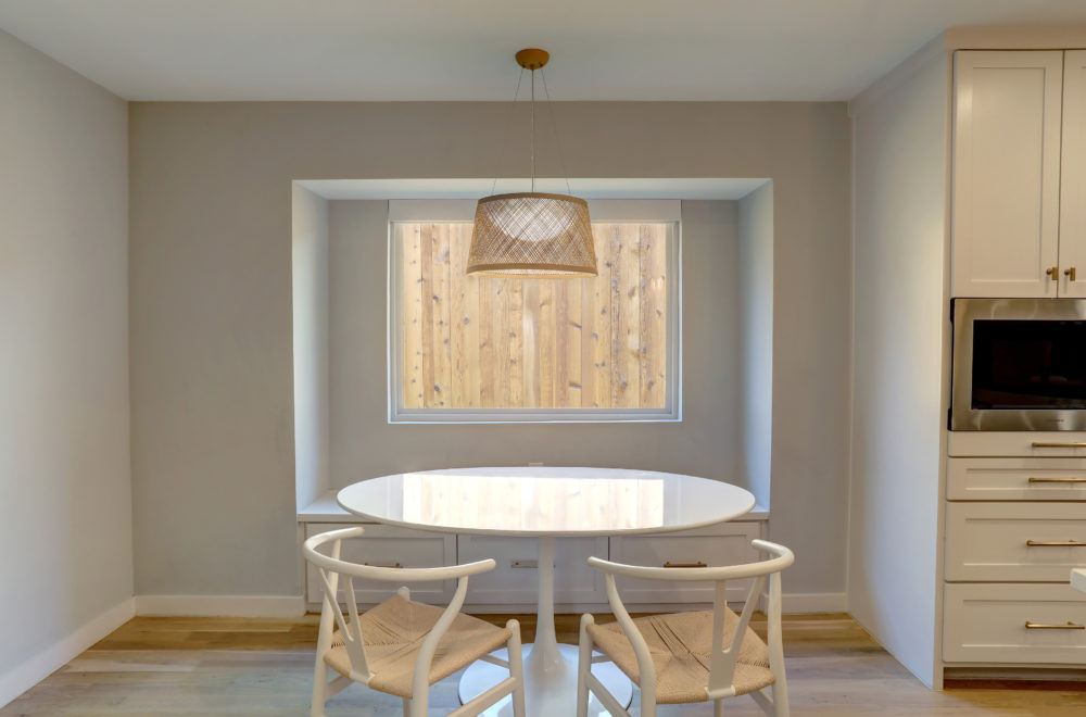 A small dining nook with a round white table and four modern chairs. The area is framed by a rectangular opening in the wall, and there is a pendant light with a rattan shade hanging above the table.