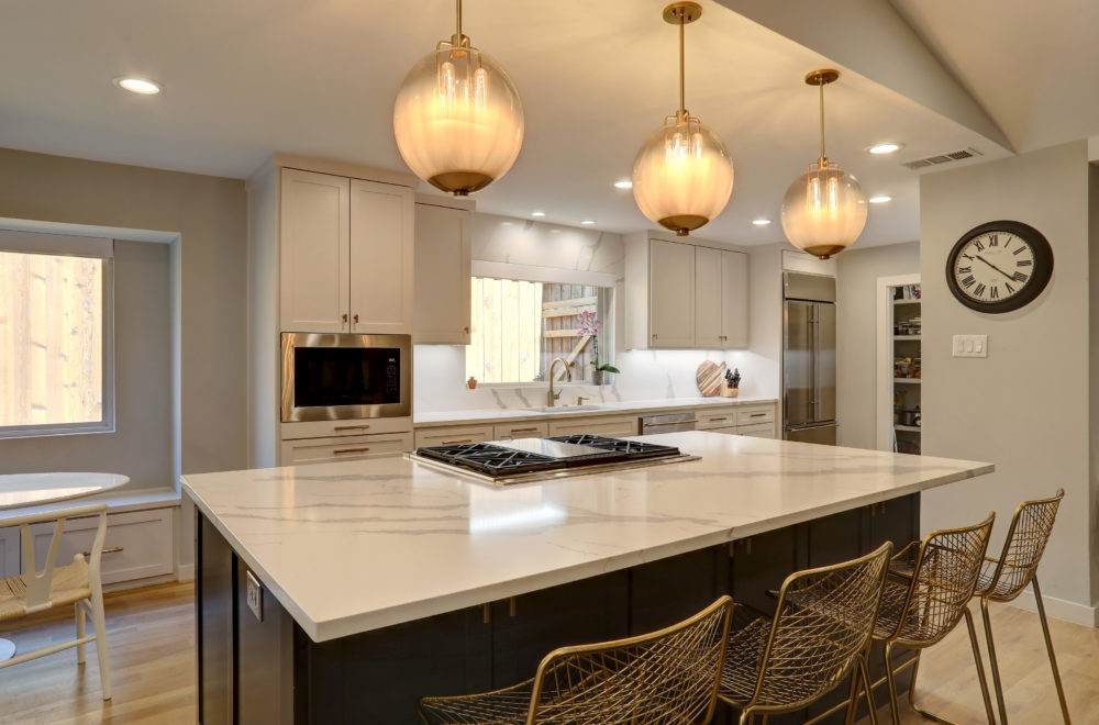     An expansive kitchen island in a modern kitchen with a white countertop, dark base cabinets, and gold-wired barstools. The kitchen has light-toned cabinetry, and there are warm globe pendant lights hanging above the island. The hardwood flooring complements the neutral color scheme of the room.