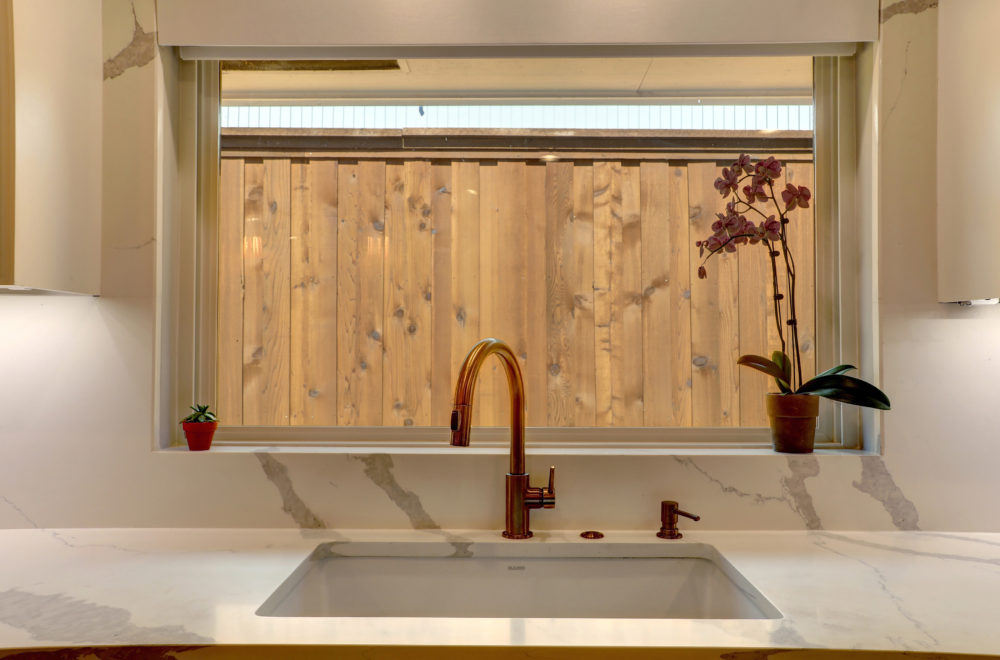 A kitchen sink area with a large window framed by wooden slats, overlooking outdoor greenery. The faucet and soap dispenser are finished in a brushed gold, contrasting with the white countertop.