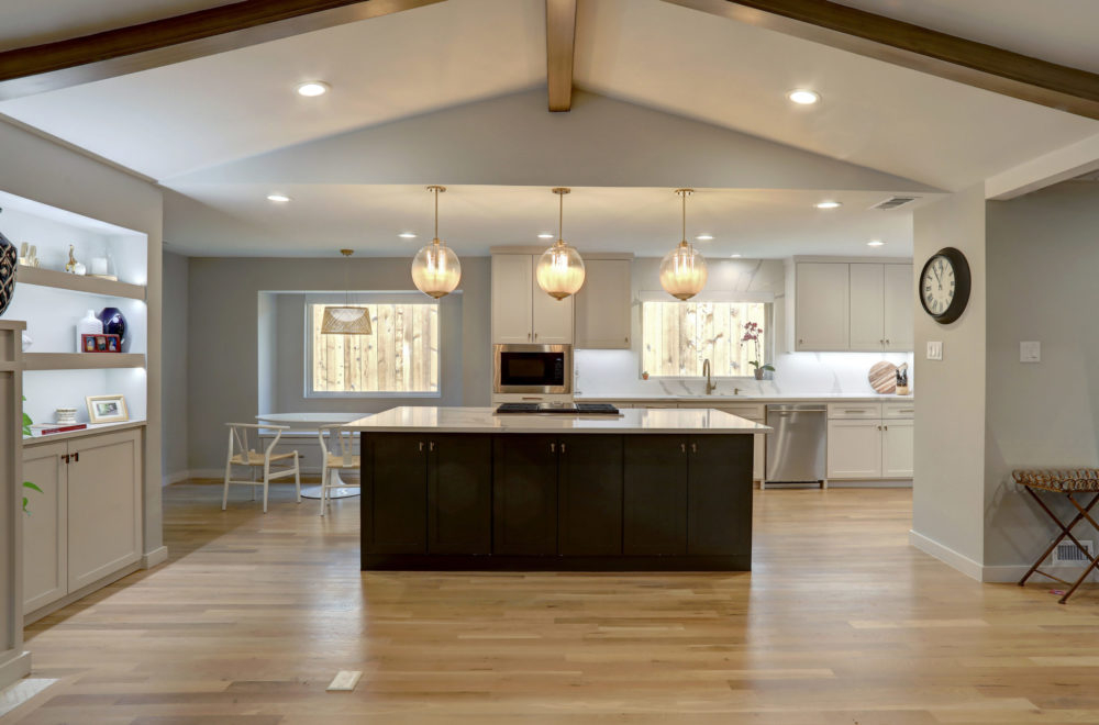 A spacious kitchen with a large central island featuring black cabinets and a light wood floor. The island has a white countertop, and there are hanging globe pendant lights above. The kitchen opens to a living area, and there's a dining space in the background.