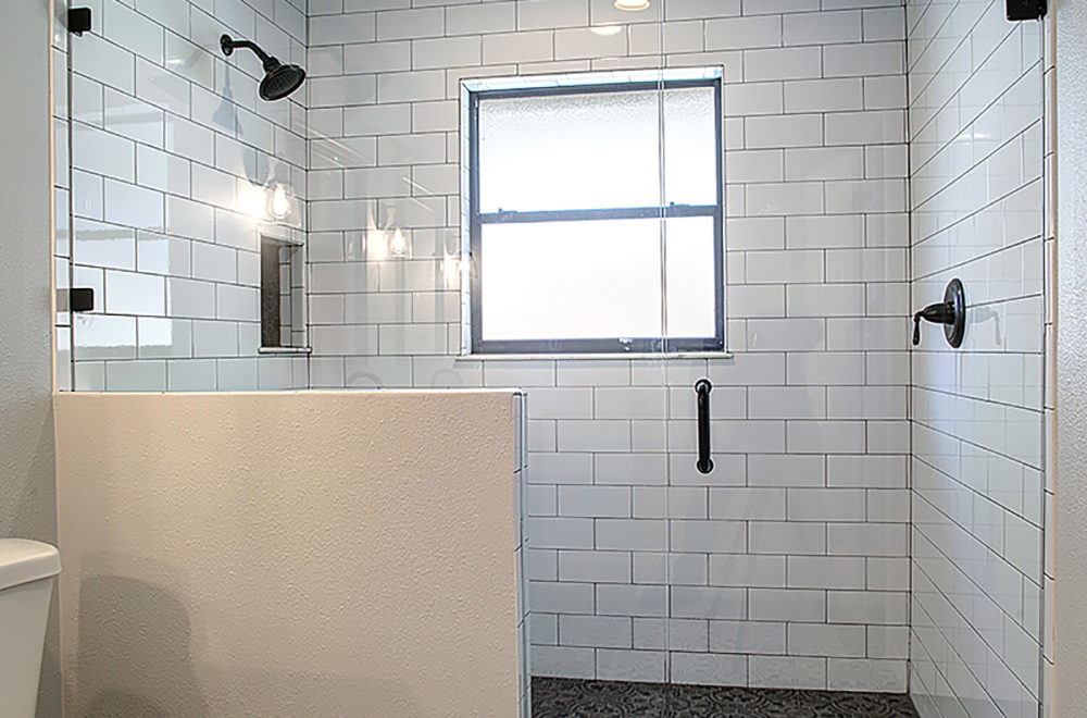 A bathroom corner showing part of a shower with a glass wall and white subway tiles. A window with frosted glass is set into the tiled wall, and there are black fixtures and a shower head visible.