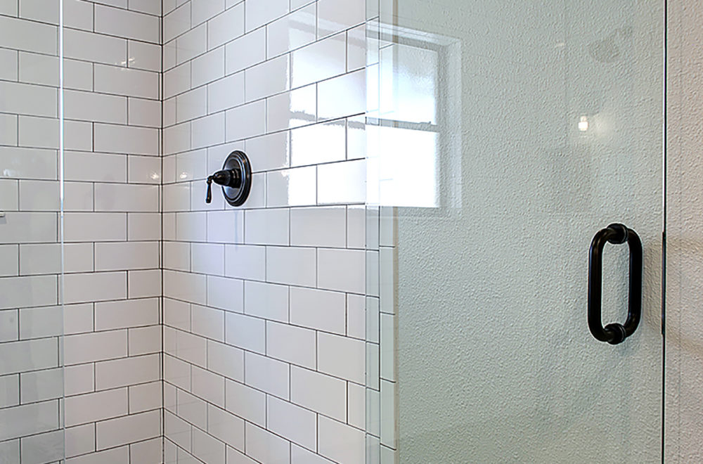 A close-up of a corner in a shower with white subway tiles and black hardware, including a shower head and control valve. The light reflects off the glossy tile surface.