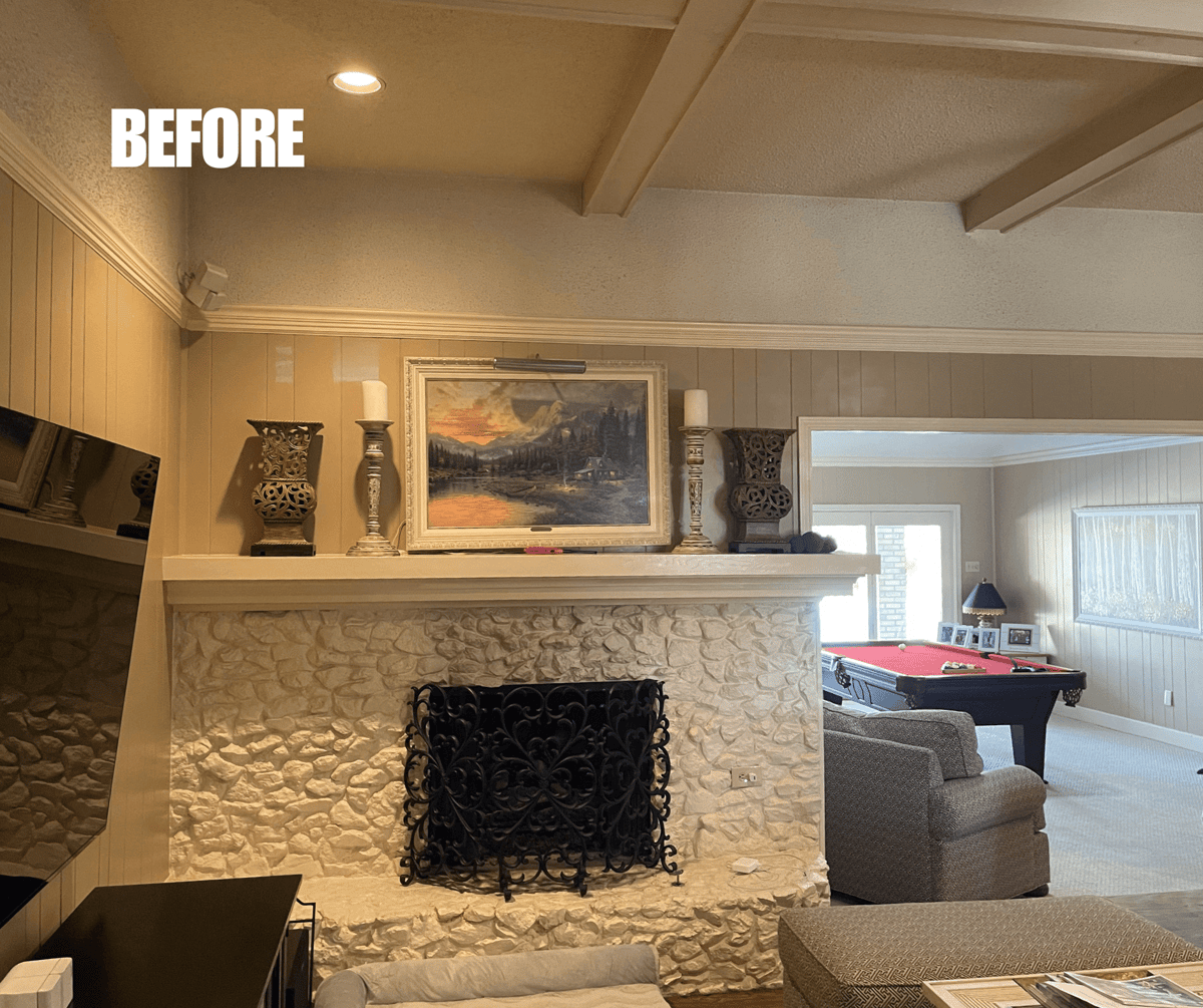 BEFORE: Living room with a stone fireplace, wall art, and decorative vases on the mantel. Pool table visible in the background.