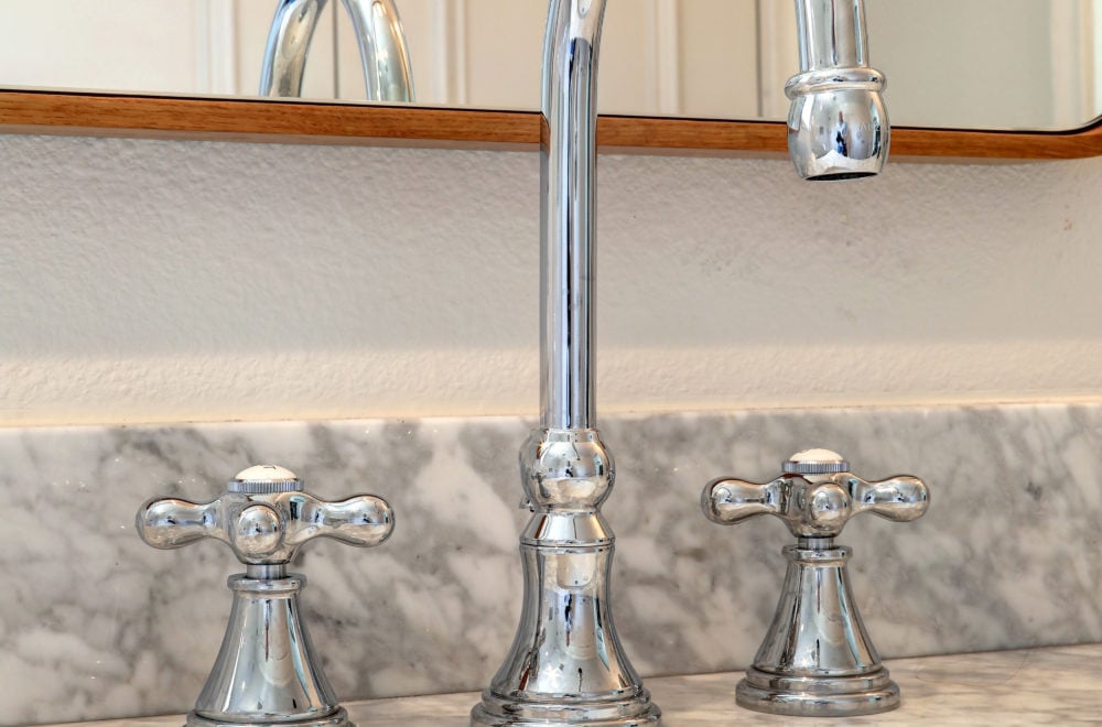 Close-up of vintage-inspired silver faucets with cross handles set against a white marble backsplash.