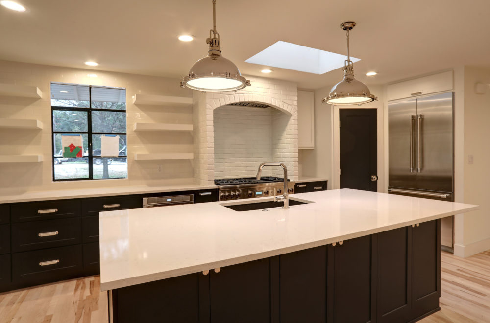 A broad view of the kitchen showing the central island with a built-in sink and a dishwasher. The kitchen has black cabinets, white countertops, and industrial-style pendant lighting. There is a large refrigerator to the right and built-in shelving in the background.