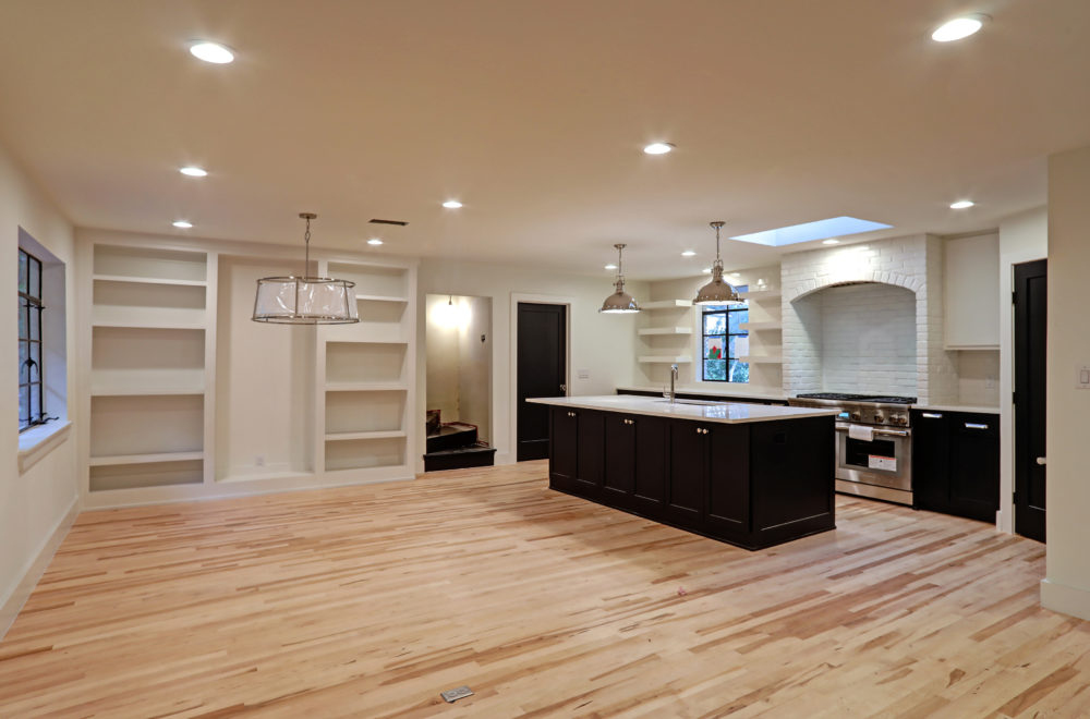 A wide view of a spacious kitchen with a central island, showing black cabinetry and white countertops. The kitchen opens to a living area with built-in shelving and recessed lighting.