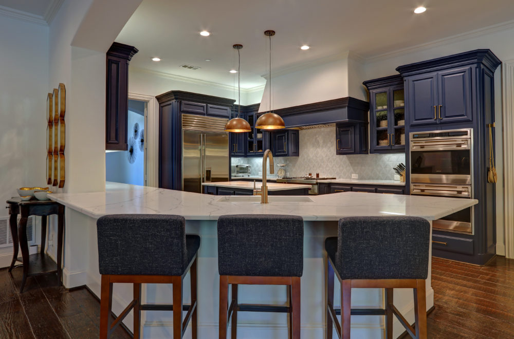 A modern kitchen with dark blue cabinets and a white countertop island. The island has bar stools with blue upholstery, and there are stainless steel appliances and pendant lights with a warm glow.
