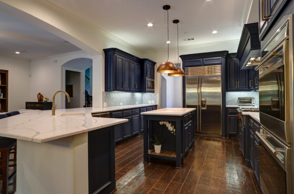 A wide view of a kitchen with a central island, featuring dark blue cabinets, white countertops, and a brass faucet. The kitchen is well-lit, with hardwood flooring and a view into an adjacent room.