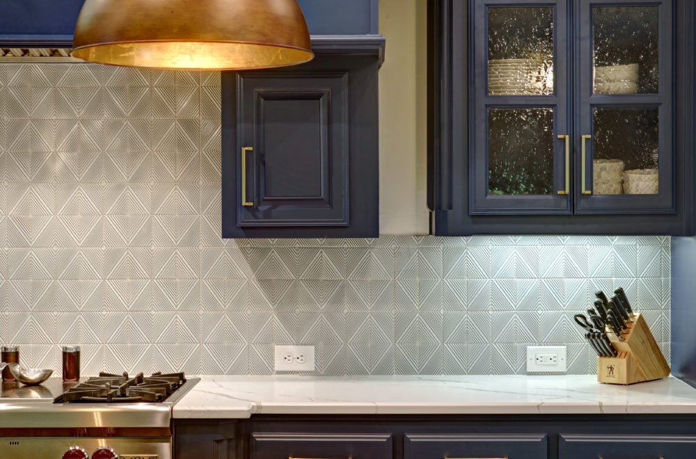 A view of a kitchen showcasing navy blue cabinets with brass hardware, a white marble countertop, and a backsplash with white geometric-patterned tiles. There are kitchen utensils in a holder and a wooden cutting board to the side.