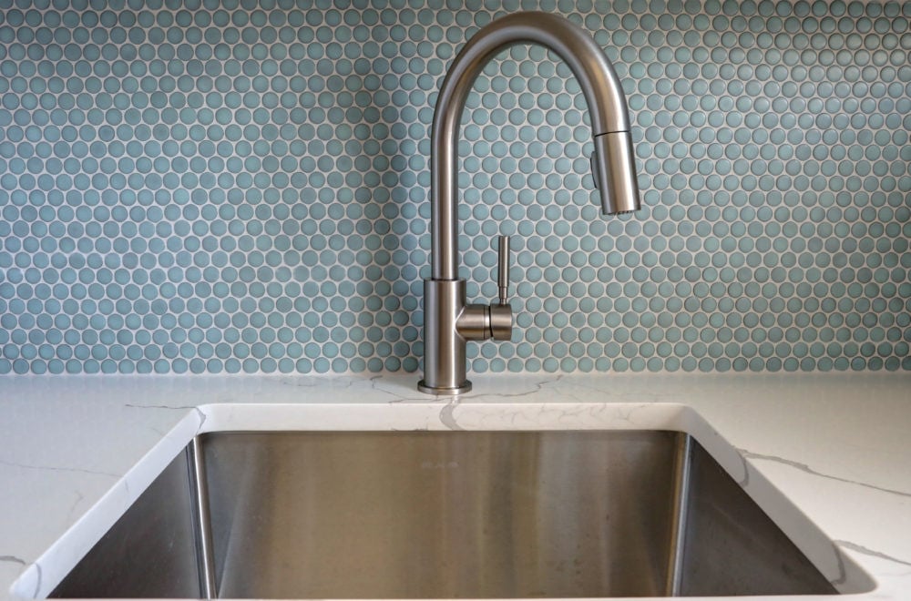 A close-up of a kitchen sink with a stainless steel finish and a modern faucet, set against a backsplash of hexagonal blue tiles.
