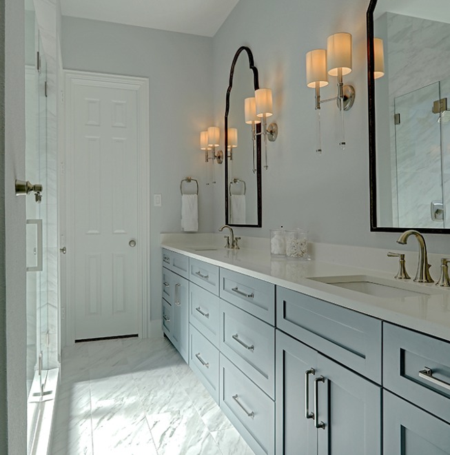 A bright bathroom with a large vanity that has white cabinets and a light marble countertop, accompanied by two sinks with gold faucets. Above are framed mirrors with sconce lighting on either side, and a white door in the background.