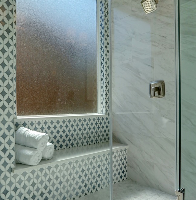 A view of a bathroom window from inside the shower, with frosted glass for privacy. The window is framed by white marble tiles with a diamond pattern accent.
