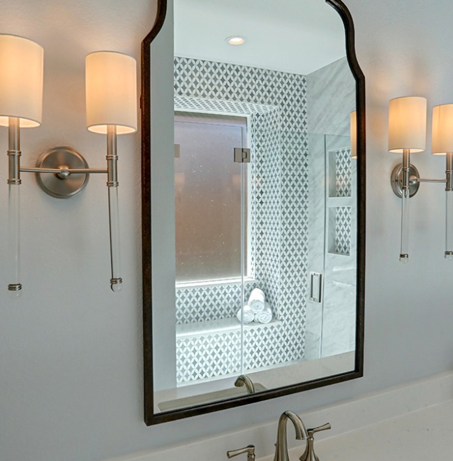 A shower with a glass door, featuring white marble tiles and an accent wall with a diamond pattern tile design. A gold shower head and control are mounted on the wall.