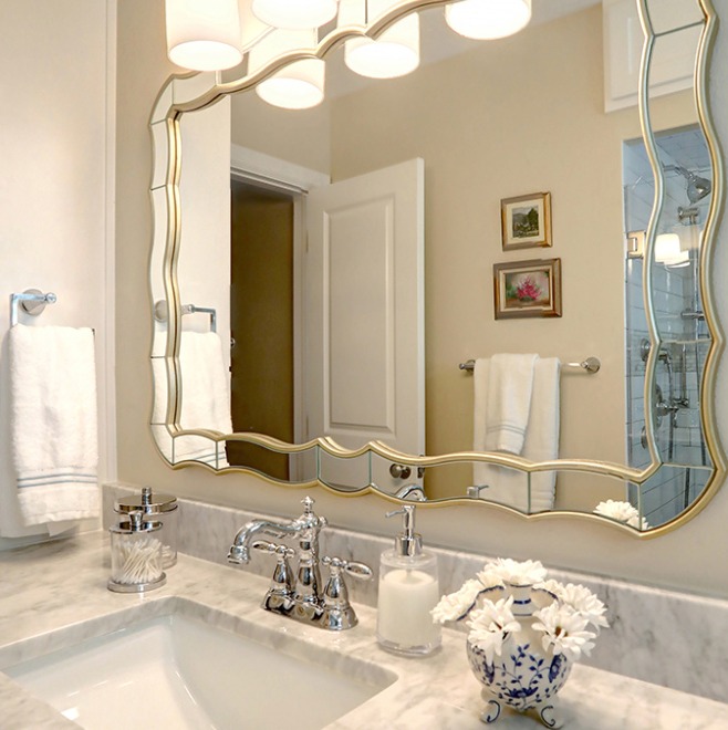 A full view of a bathroom with a large vanity mirror with a distinctive curved frame reflecting the tiled wall and a towel hanging on a rack. The room has a marble countertop with a sink and chrome fixtures, and there is a framed picture hanging on the wall.