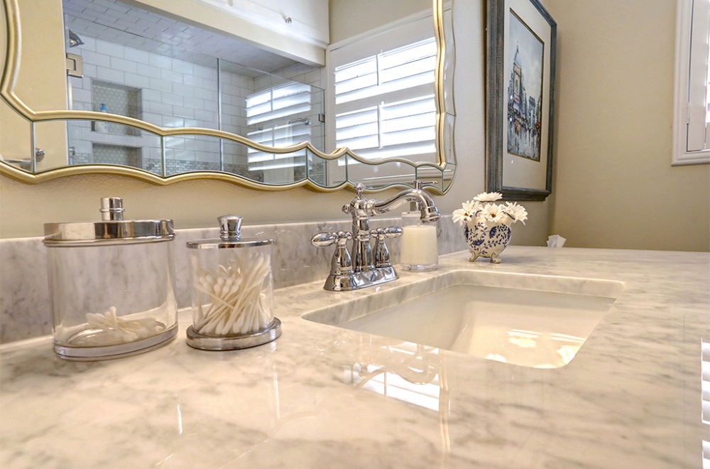 A bathroom countertop displaying various accessories such as a soap dish, toothbrush holder, and containers for cotton balls. The countertop is marble, and there is a large mirror with a unique wavy frame above it.