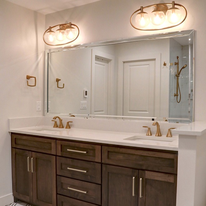 A double sink bathroom vanity with dark wood cabinetry and a white countertop. Above the vanity, there is a large mirror reflecting a glass shower enclosure. The fixtures, including the faucet and towel ring, are finished in brushed gold.
