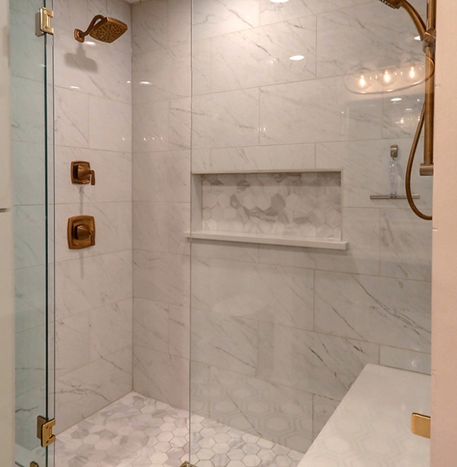 A shower corner with white marble tiles and a built-in niche for toiletries. The shower fixtures, including a rainfall showerhead and a hand-held sprayer, are finished in brushed gold, matching the niche's trim.