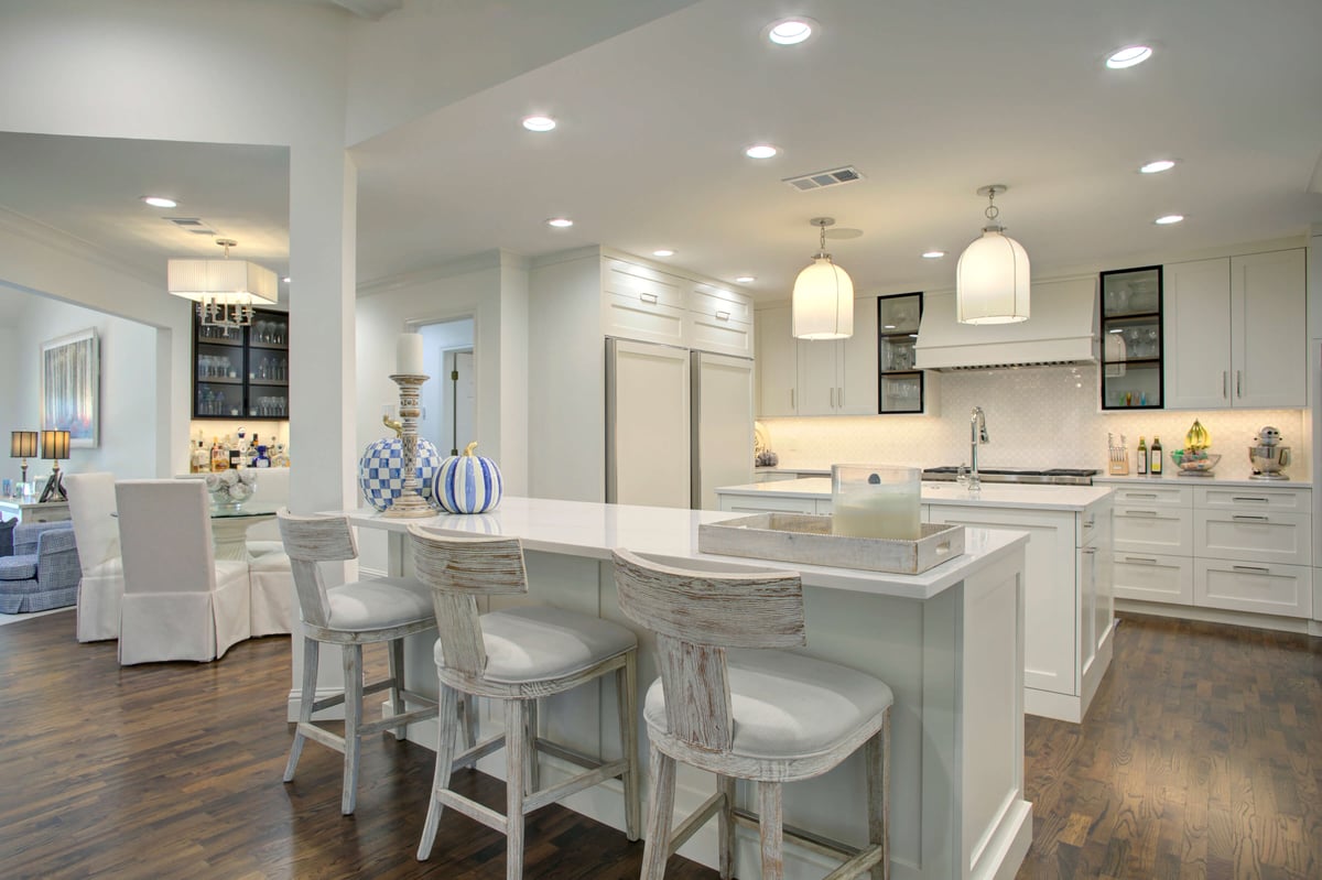 Modern kitchen with white cabinetry, quartz countertops, and kitchen island. Pendant lights hang above, and a dining area is visible in the background.