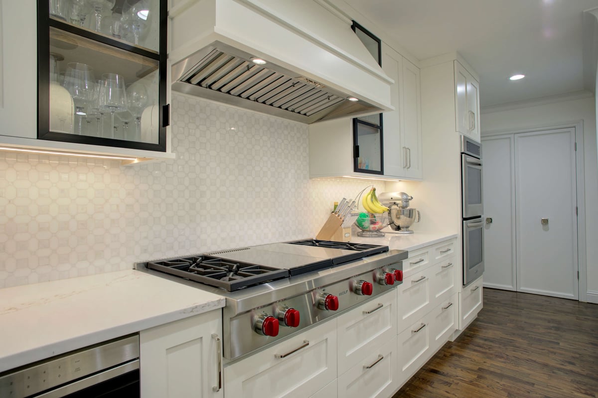 Modern kitchen with a stainless steel Wolfe cooktop, white cabinetry, a hexagonal tile backsplash, and dark wood flooring. Countertop holds a fruit bowl and a stand mixer.