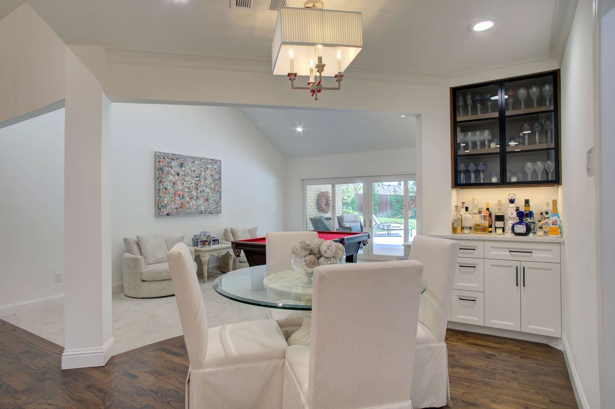 Dining area with a round glass table and four white chairs. A bar with shelves and glassware is on the right. In the background, a living area with a pool table and seating is visible.