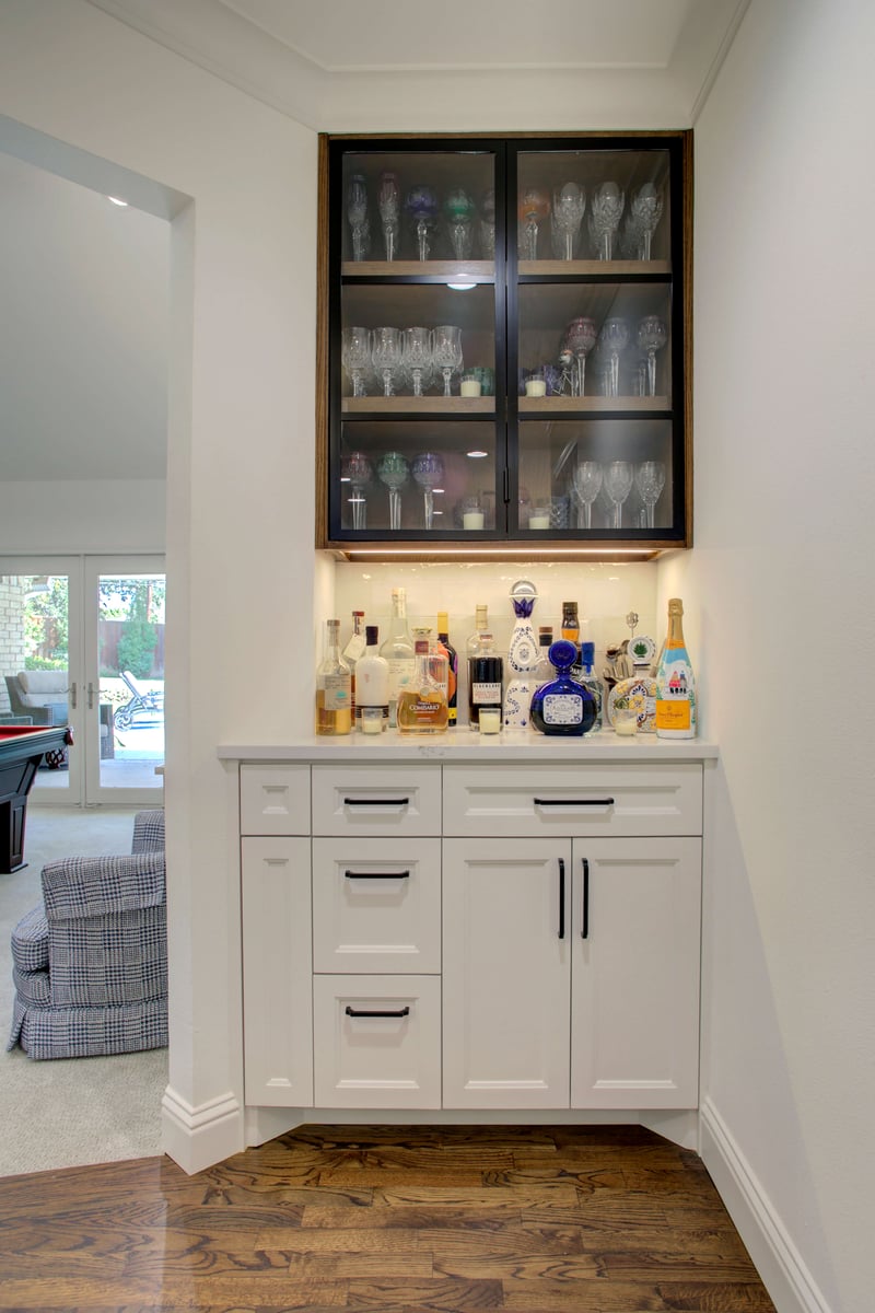 Home beverage bar with white cabinets, various liquor bottles on the counter, and glassware displayed in an upper cabinet with glass doors.