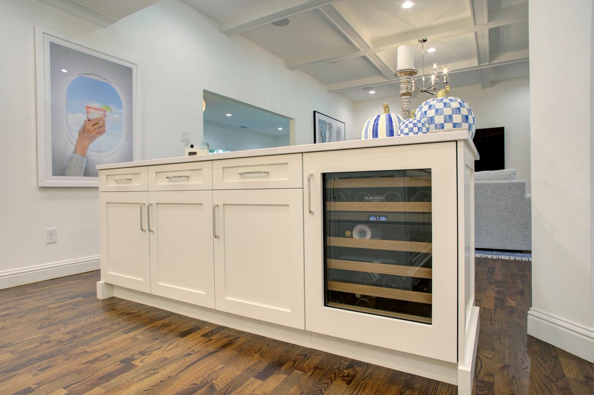 A white kitchen island with four drawers and a wine cooler below. A blue and white decorative item is on top, with a framed picture on the wall in the background.