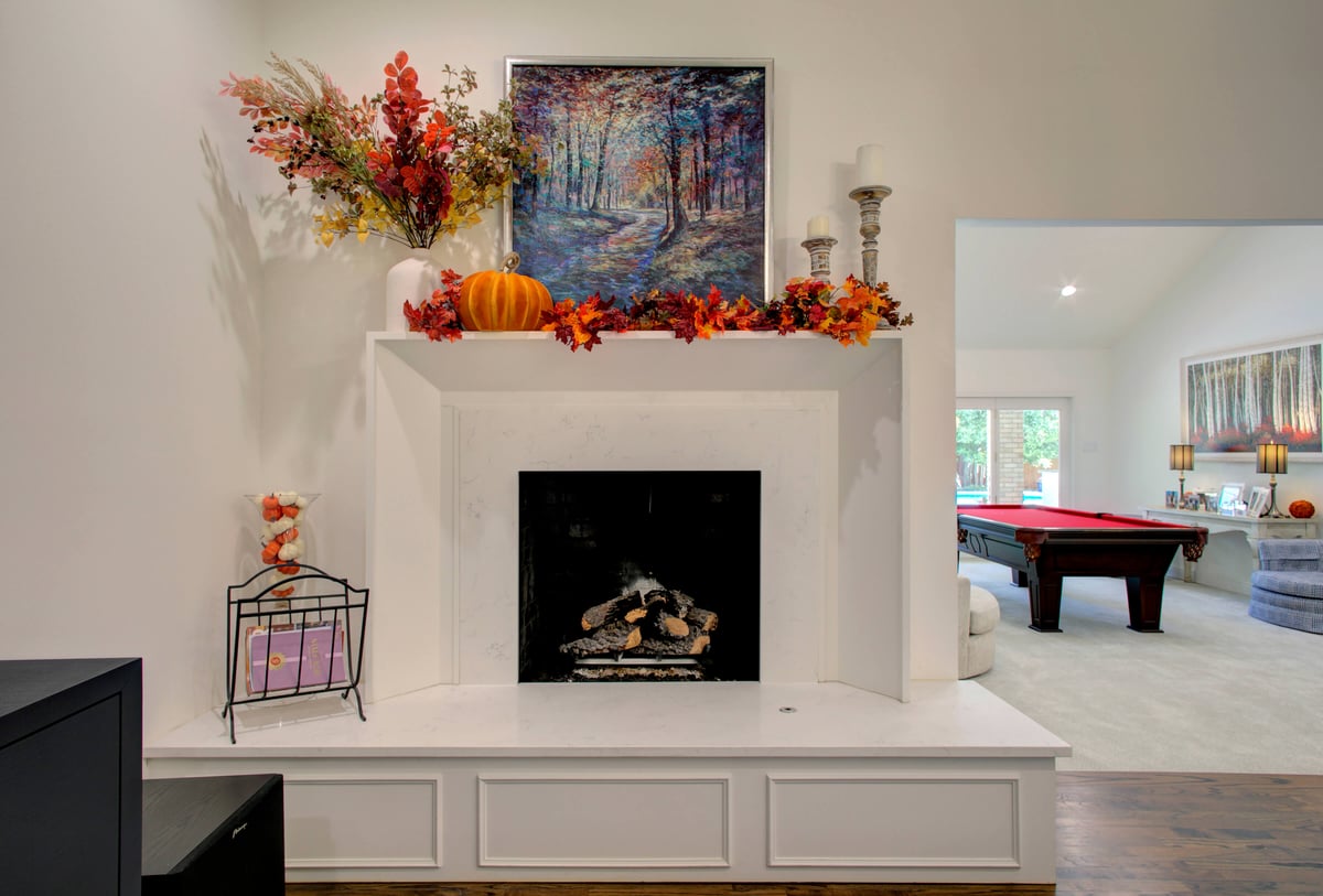 A fireplace decorated with autumn leaves, a pumpkin, and candles. A painting hangs above. In the background, a room with a red pool table is visible.