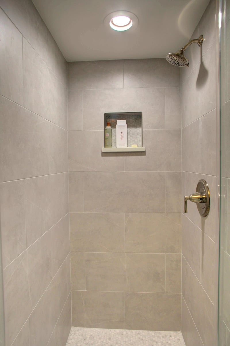 A modern shower with large profile tile walls, a metallic showerhead, and a built-in shelf holding toiletries.