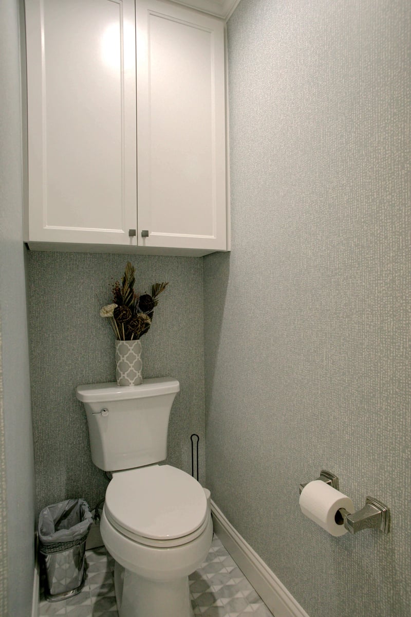 A small bathroom with a white toilet, wall-mounted cabinet, floral arrangement on the tank, toilet paper on a holder, and a trash bin. The walls are covered with textured wallpaper.