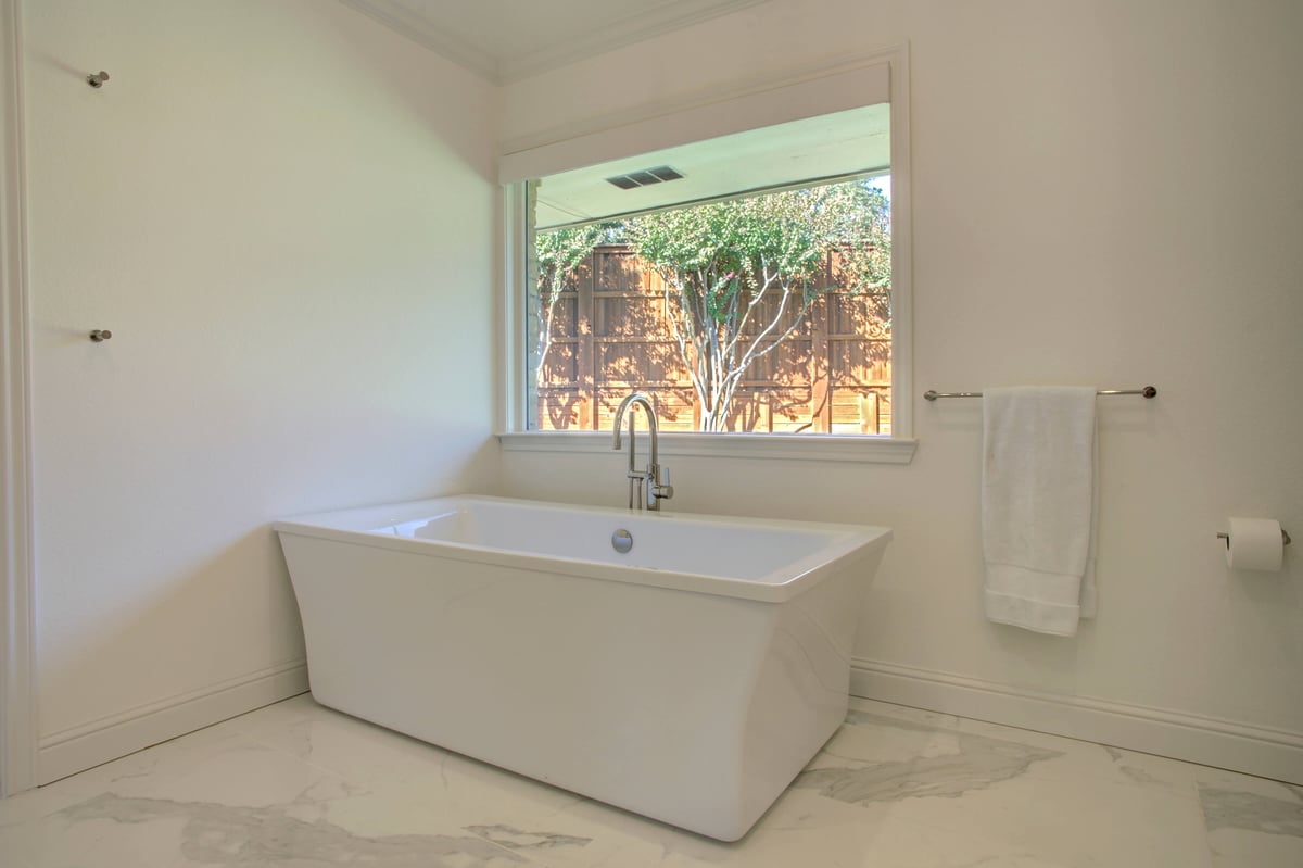 Minimalist bathroom with a white freestanding bathtub, large window with outdoor view, and towel rack on the wall. Located in North Dallas