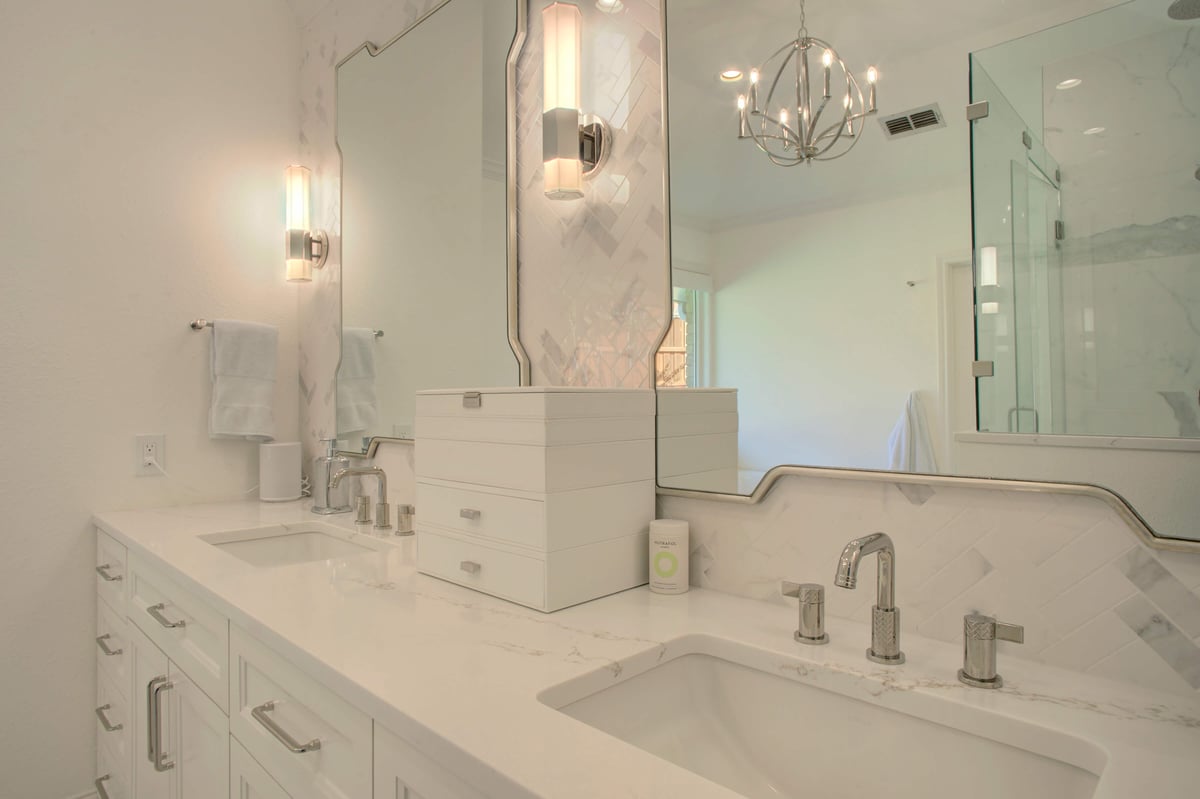 Modern bathroom with dual sinks, white countertops, silver faucets, and illuminated mirrors. A glass shower enclosure is visible in the background, along with a decorative chandelier.