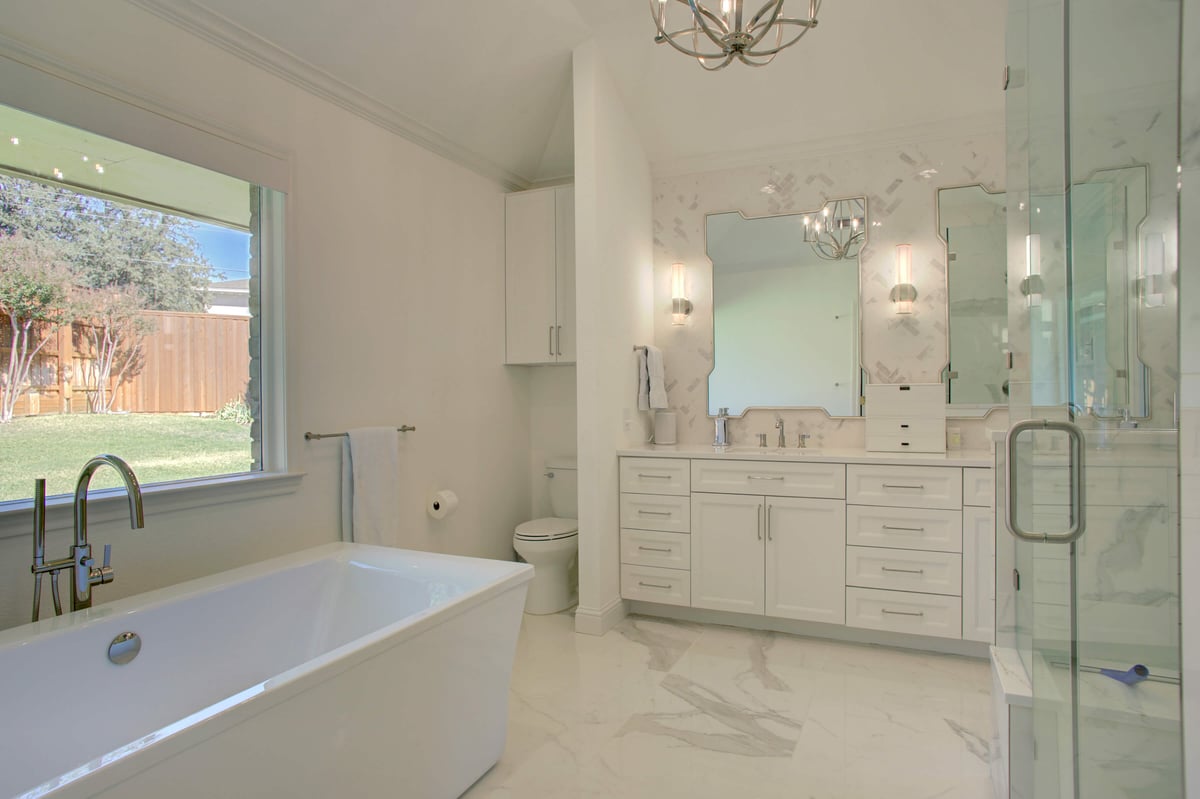 A modern bathroom with a white bathtub, dual-sink vanity with mirrors, and a glass-enclosed shower. A window offers a view of a fenced backyard.