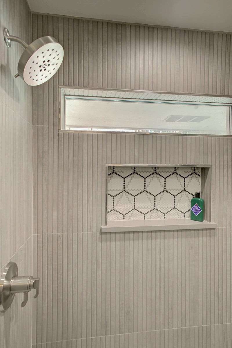 Shower with a modern design, featuring a large showerhead, gray vertical tiles, a small window, and a recessed shelf with hexagonal tiles holding a bottle.