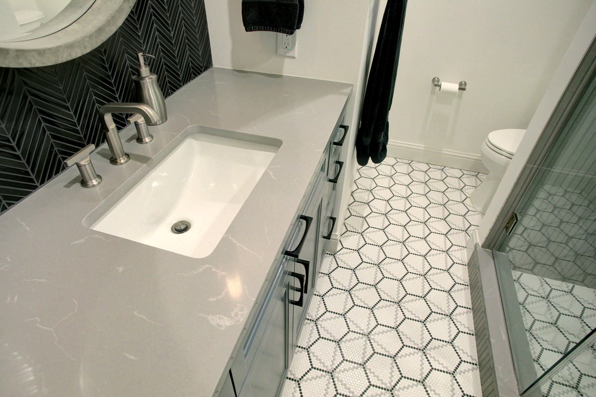 Modern bathroom with a gray countertop, rectangular sink, and herringbone-patterned backsplash. The floor features hexagonal tiles, and a glass shower is visible on the right.