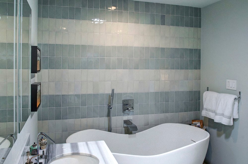 A view of a modern bathroom with a double vanity, large mirrors, and a walk-in shower with glass doors. The room is tiled with a combination of large stone tiles and smaller accent tiles.