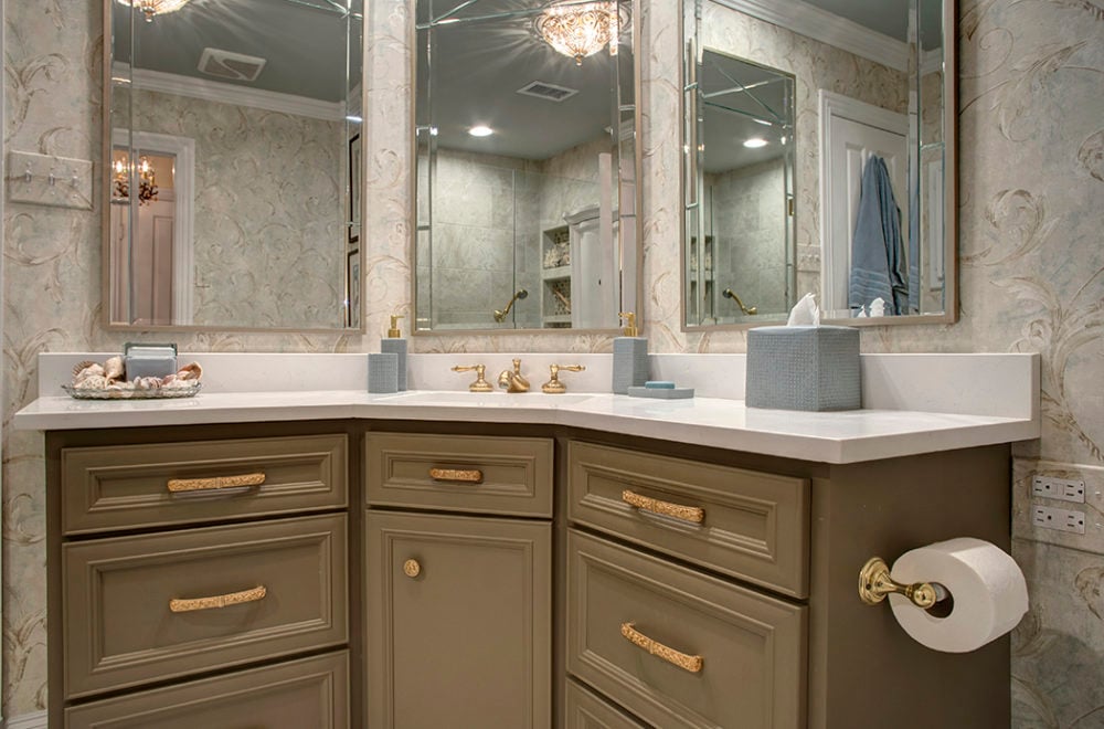 A classic bathroom vanity with taupe cabinetry and gold hardware. The countertop is light-colored, and there is a large mirror with decorative molding. The walls are adorned with a patterned wallpaper.