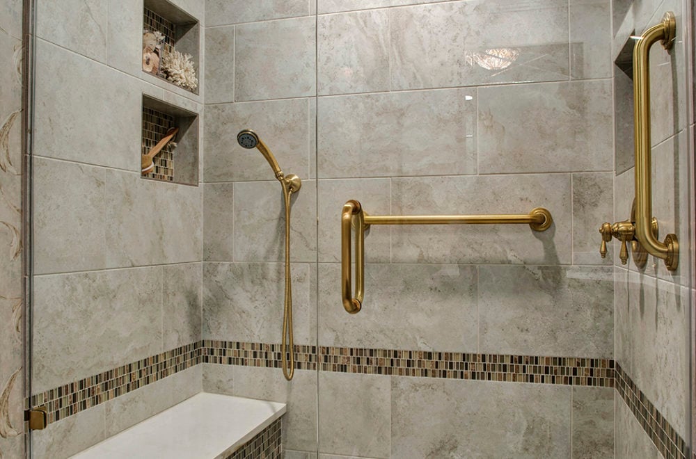 A close-up of a bathroom corner with marble walls and a small mosaic tile accent. There is a gold grab bar and fixtures, adding a touch of elegance.