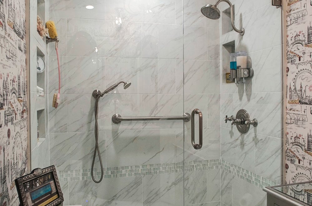 A corner view of a marble-tiled shower with glass doors. The shower fixtures are mounted on the marble walls, and there is a built-in niche for toiletries.