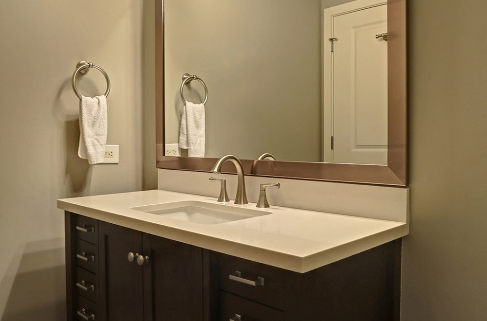 A modern bathroom vanity with dark wood cabinets and a light countertop with a single sink. Above it, there's a large mirror flanked by two wall-mounted lights. Hand towels hang on circular holders on the wall.