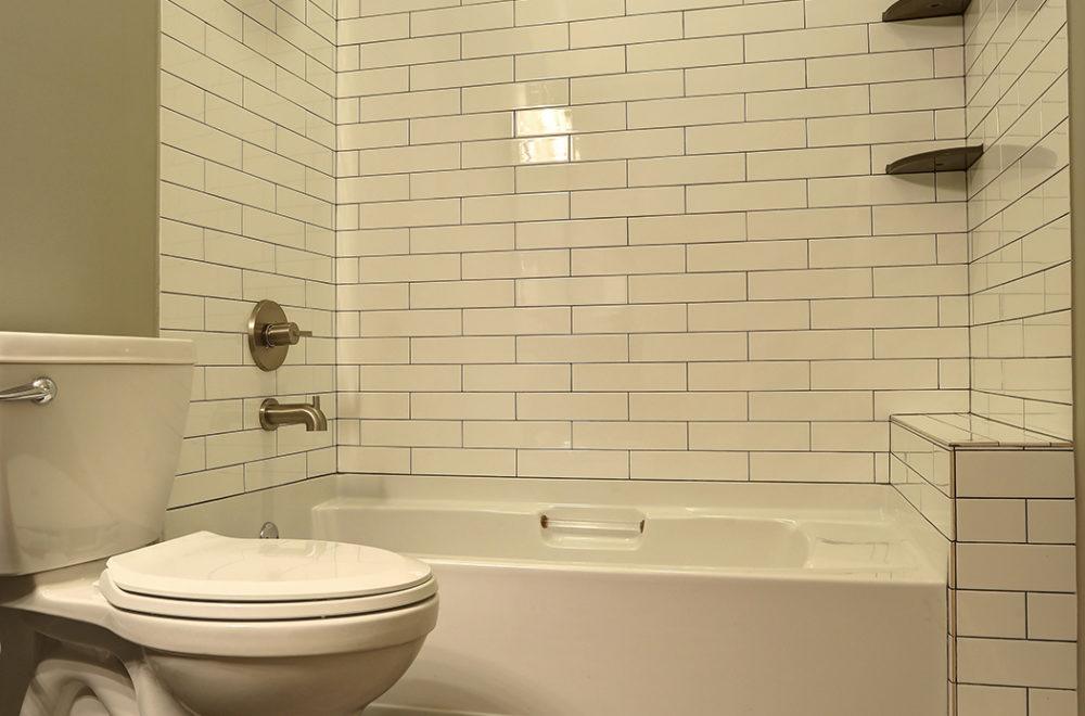 A bathroom corner with a toilet beside a bathtub, both with a beige subway tile wall behind them. There's a small built-in shelf within the tiling above the bathtub, and the floor is tiled with gray wood-look tiles.