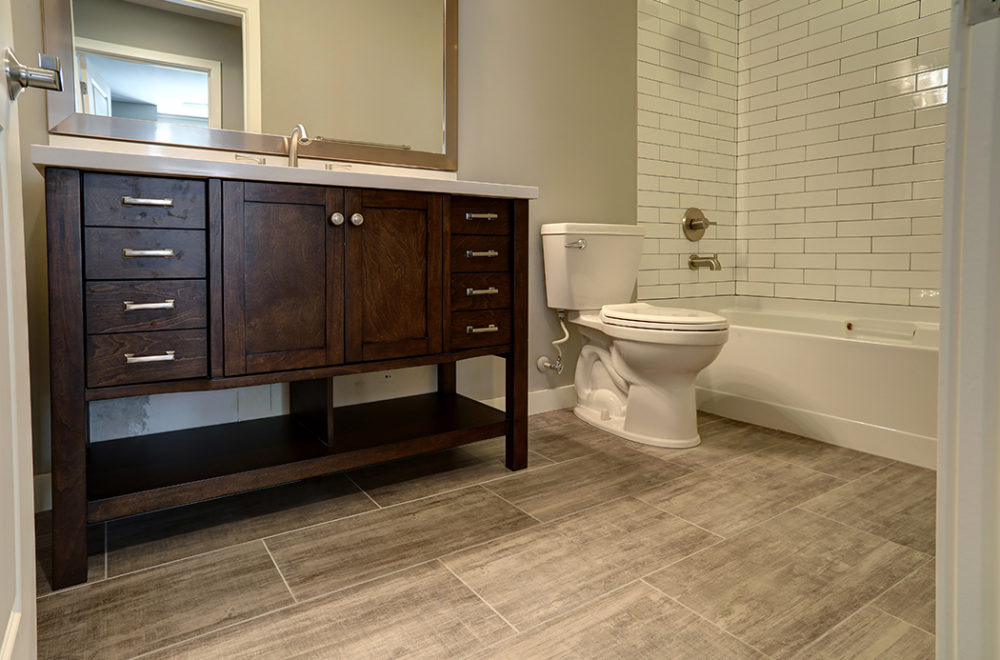 A spacious bathroom vanity with dark wood and an open shelf below. It has a light countertop with a single sink and a large rectangular mirror above it. The flooring is gray wood-look tile, and there is a toilet to the right.