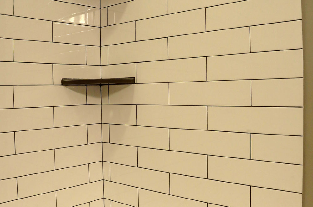 An interior of a shower with beige subway tiles on the walls, featuring a dark brown shower shelf installed within the tiling. The corner shows the tile work wrapping around.