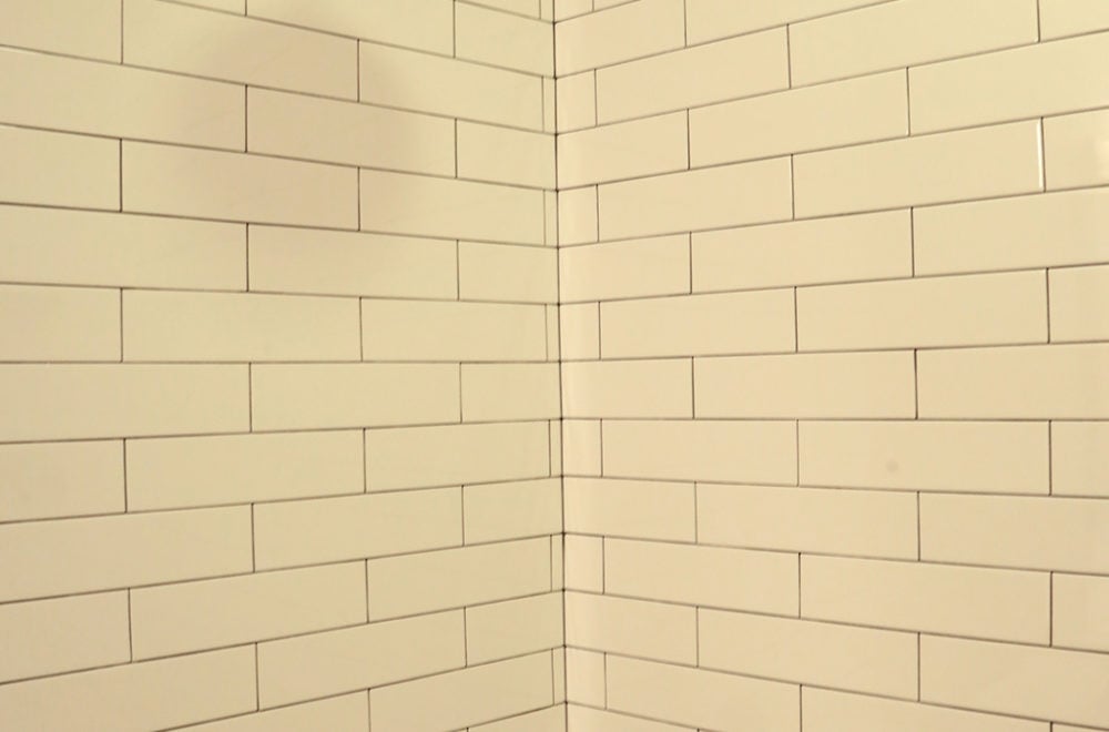 A corner view of a tiled bathroom showing the continuous subway tile pattern wrapping around two walls. The corner edge is neatly finished, and the tiles are a light beige color.